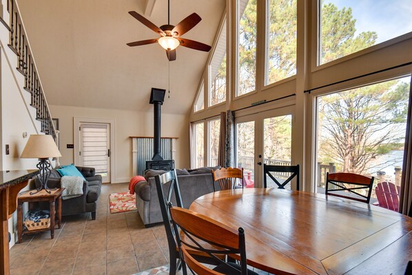A spacious living area with a large wooden dining table, comfortable seating, a fireplace, and large windows offering a view of trees.