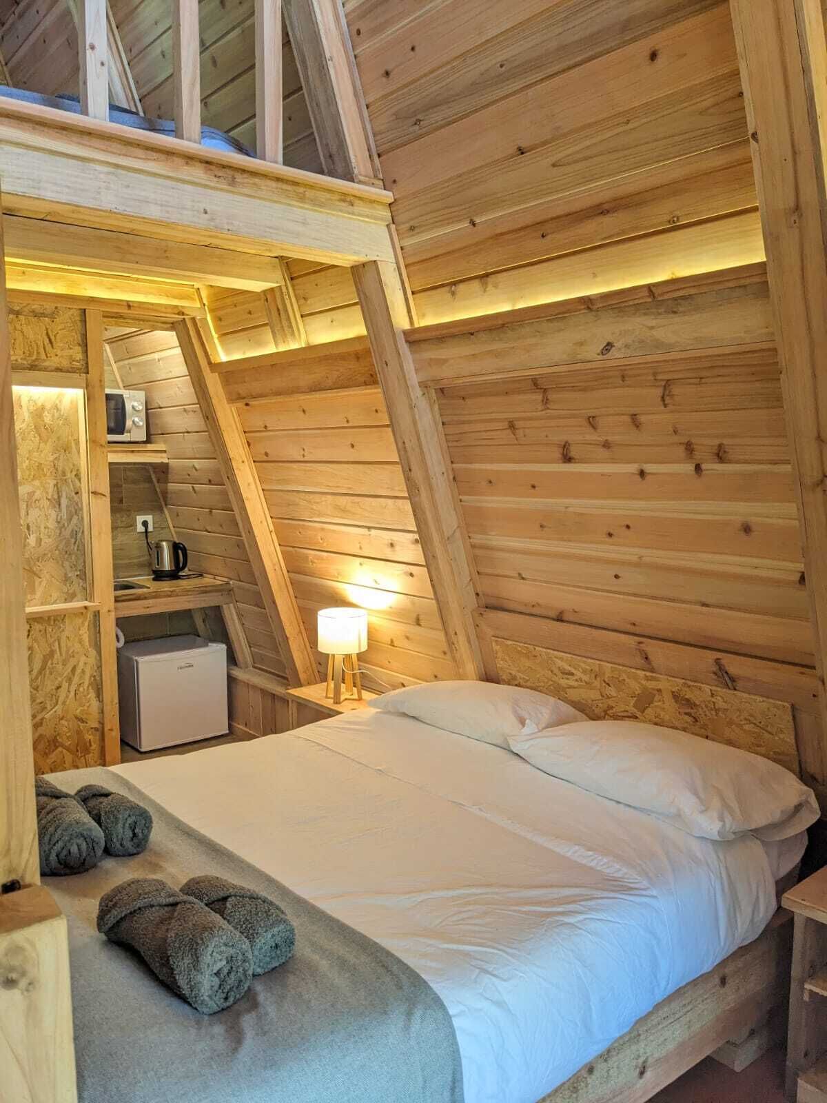 Un dormitorio acogedor en el ático con techo inclinado de madera, una cama con ropa blanca, una mesita de noche con lámpara y una kitchenette pequeña con microondas y cafetera.