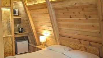Un dormitorio acogedor en el ático con techo inclinado de madera, una cama con ropa blanca, una mesita de noche con lámpara y una kitchenette pequeña con microondas y cafetera.