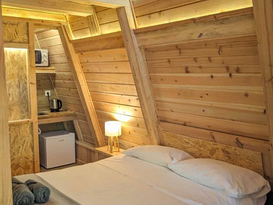 Un dormitorio acogedor en el ático con techo inclinado de madera, una cama con ropa blanca, una mesita de noche con lámpara y una kitchenette pequeña con microondas y cafetera.