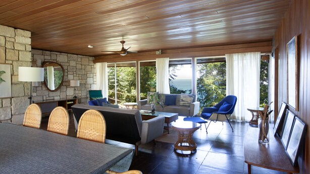 A spacious room with a stone wall, wooden ceiling, and large windows. There is a dining table with chairs, a sofa, and a blue chair. - Luxury Three Bedroom Sea View Villa, Frenchman's Cove Resort (Drapers)