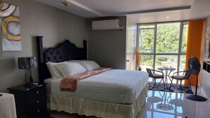 A bedroom with a bed, bedside table, lamp, and a view of the outdoors through a large window. - Condo Serenity (Portmore)