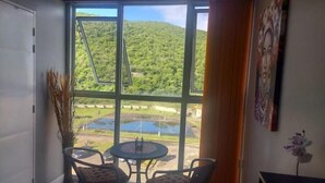 A room with a large window, a small table with chairs, a potted plant, and a painting on the wall. - Condo Serenity (Portmore)