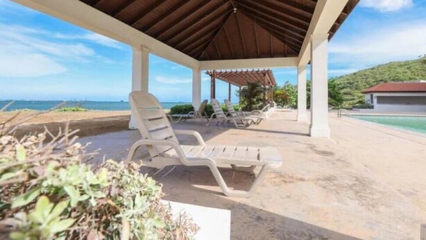 A covered outdoor seating area with lounge chairs, a pool, and a view of the ocean. - Condo Serenity (Portmore)