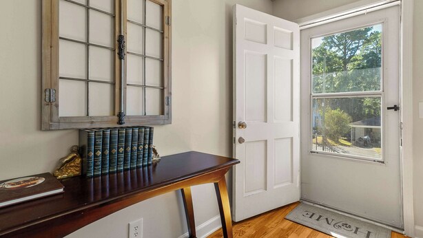 A room with a wooden door, a window with a view of greenery, a wooden table with books, and a decorative mirror. - Comfort Cottage (Northport)