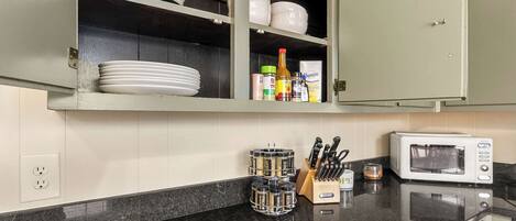 Uma cozinha com bancada de granito preto, micro-ondas branco, suporte para facas e prateleira para temperos.