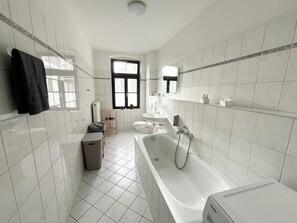 Une salle de bain moderne équipée d’une baignoire, de toilettes et d’un lavabo. Une fenêtre laisse entrer la lumière naturelle, et une étagère contient des articles de toilette.