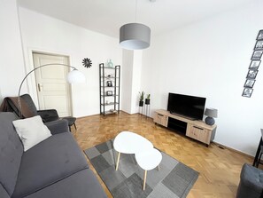 Un salon moderne avec un canapé gris, une table basse blanche, un téléviseur posé sur un meuble en bois, une lampe sur pied et une bibliothèque.