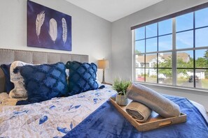Ein Schlafzimmer mit großem Fenster, einem Bett mit blau-weißer Bettwäsche, einem Federbild und einem Holftablett mit Handtüchern.