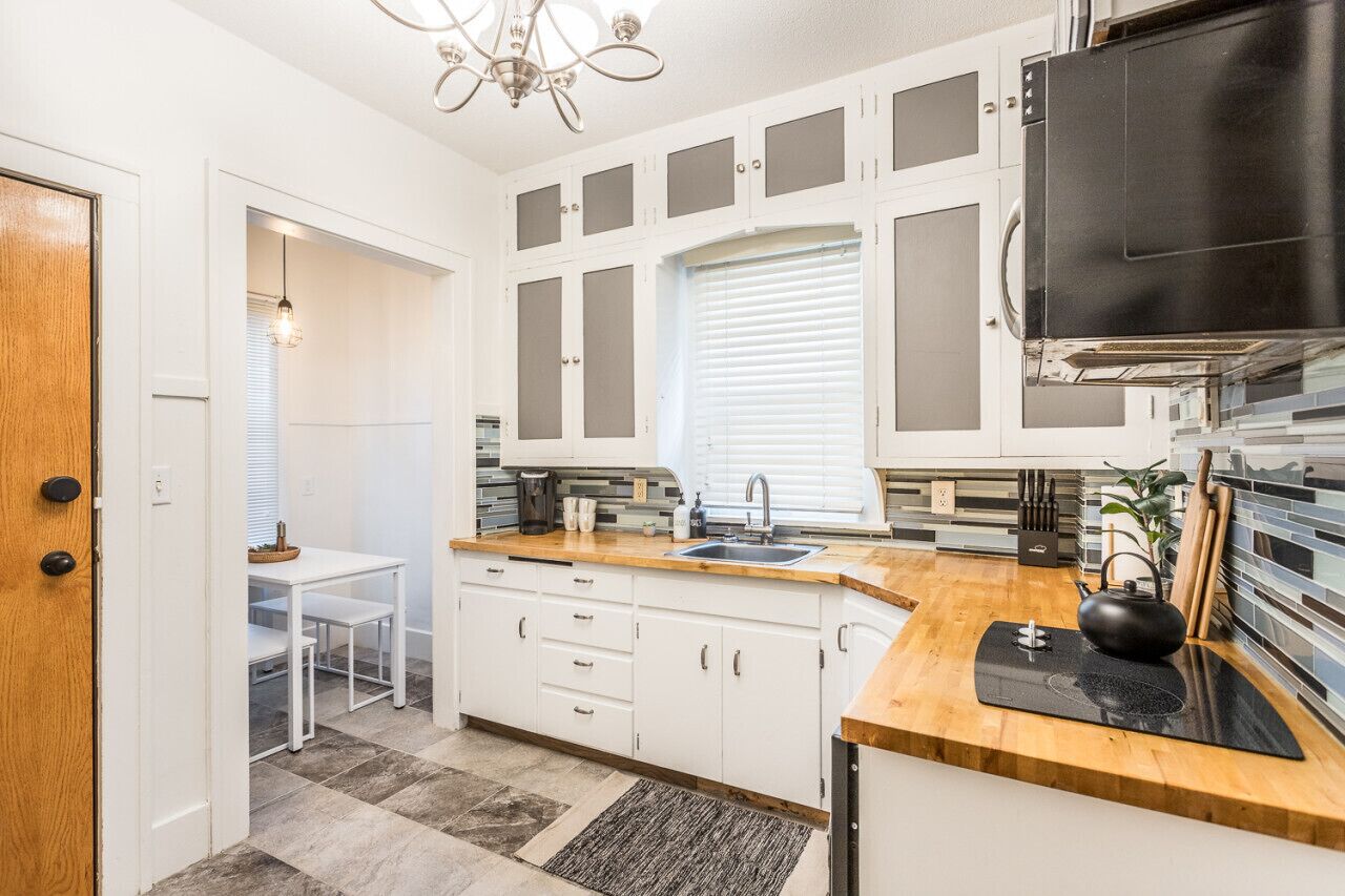 A kitchen with white cabinets, a wooden countertop, a black kettle, and a window with blinds.