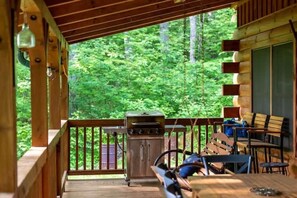 Une terrasse en bois avec un barbecue et des chaises, donnant sur une zone boisée.