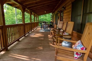 Eine überdachte Veranda mit Holzsesseln und -tischen, Blick auf den Wald und ein rustikales Blockhaus.