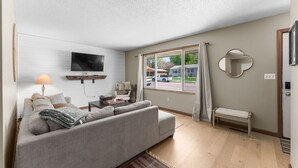 Ett vardagsrum med en soffa, en tv, en lampa, ett fönster med gardiner och en spegel.