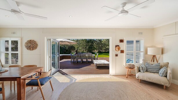 A spacious living room with a wooden dining table, a sofa, and a view of an outdoor deck with a table and chairs. - MacMasters Malibu - Lakefront by HolidayCo. (MACMASTERS BEACH)