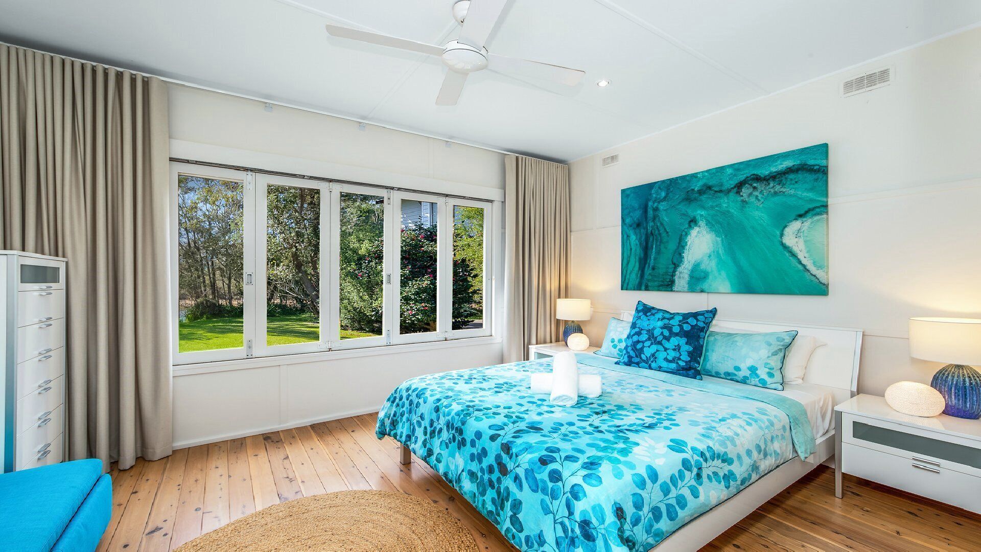 A bedroom with a large window, a bed with blue floral bedding, a ceiling fan, and a painting of a wave.