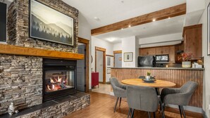 A modern kitchen with a stone fireplace, wooden beams, and a dining area with grey chairs. - Red Maple Retreat | Hot Tub | Pool | Gas Fireplace (Radium Hot Springs)