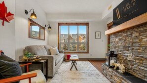 A cozy living room with a stone fireplace, a large window offering a view of buildings, a grey sofa, and a red maple leaf decoration. - Red Maple Retreat | Hot Tub | Pool | Gas Fireplace (Radium Hot Springs)