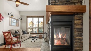 A cozy living room with a stone fireplace, a red chair, a sofa, and a large window offering a view of houses. - Red Maple Retreat | Hot Tub | Pool | Gas Fireplace (Radium Hot Springs)