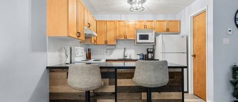 A kitchen with wooden cabinets, a microwave, a refrigerator, and two bar stools.
