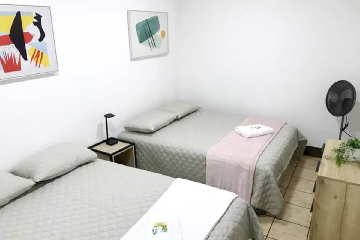 A small, clean room with two beds, a bedside table, a fan, and artwork on the walls.