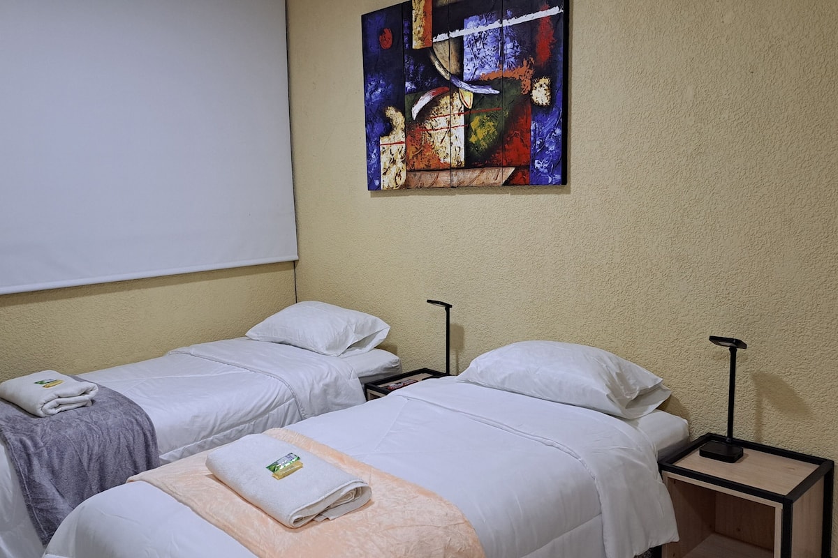 A hotel room with two single beds, white bedding, a small bedside table with a lamp, and a large abstract painting on the wall.
