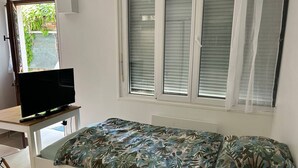Ett sovrum med en säng, en tv på ett tv-bord, ett fönster med persienner och utsikt över grönområden utanför.
