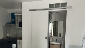 Ett kompakt bostadsutrymme med ett matbord i trä, en tv, en vit stol, ett köksområde med skåp, en diskho och en tvättmaskin.