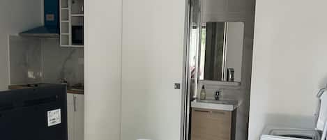 A compact living space with a wooden dining table, a TV, a white chair, a kitchen area with cabinets, a sink, and a washing machine.