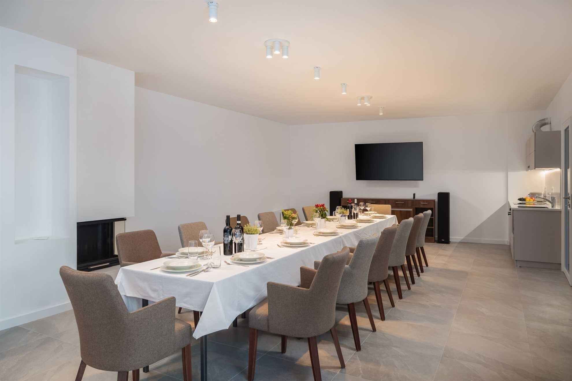 A modern dining room with a long table set for a meal, a flat-screen TV mounted on the wall, and a kitchen area with a refrigerator and sink.