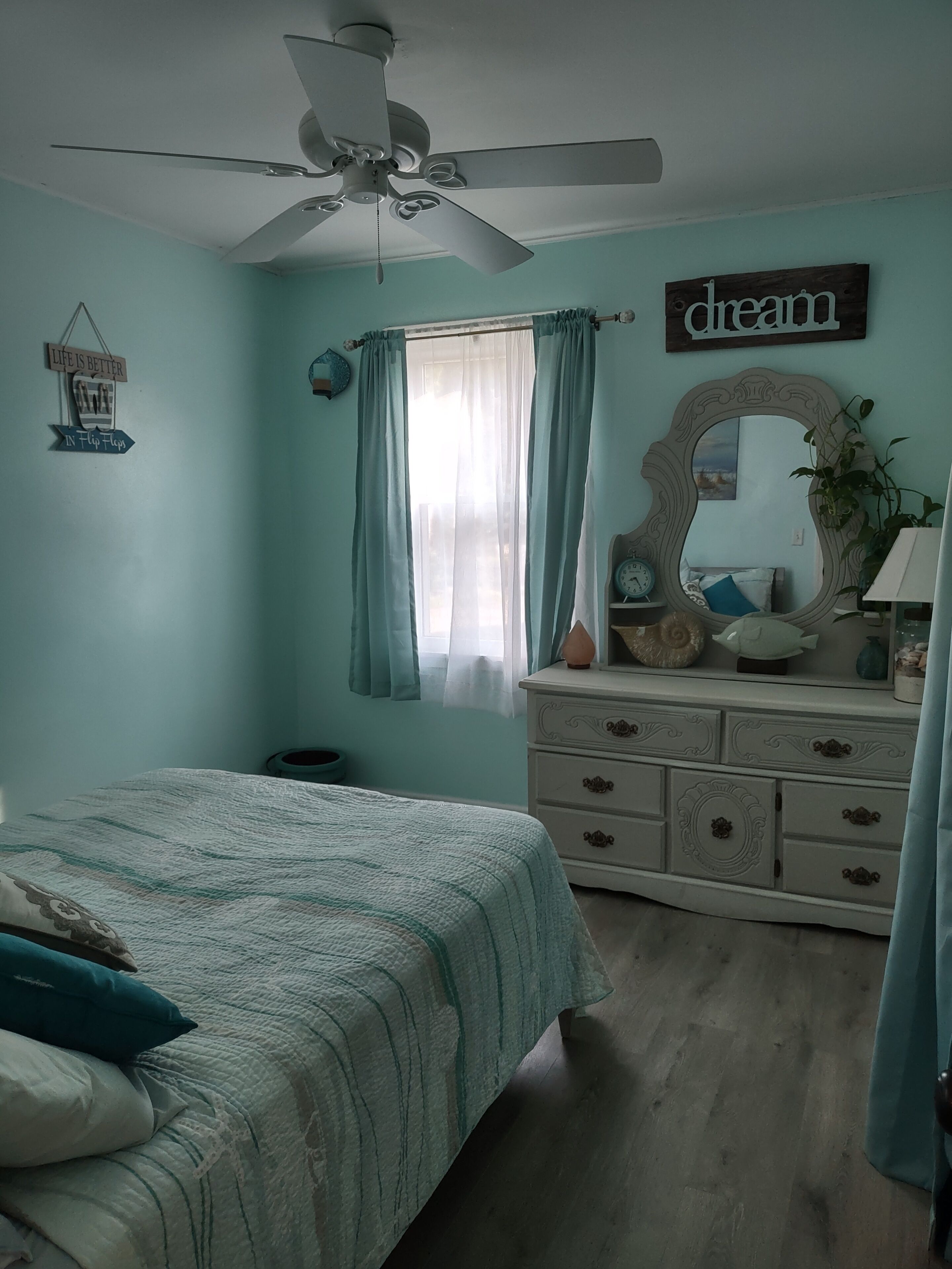A bedroom with a bed, a dresser, a ceiling fan, a window with curtains, and a wall with a mirror and a sign.