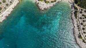 Ett flygbildsbild av ett kustområde med ett klart turkosfärgat hav, en strand med människor och en byggnad med ett rött tak.