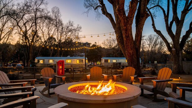 A fire pit surrounded by Adirondack chairs, with a row of RVs parked in the background.