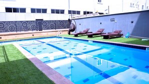 A rectangular swimming pool with a tiled pattern, surrounded by a grassy area and a patterned wall.