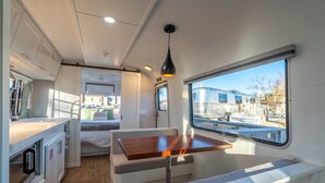 A compact RV interior with a kitchenette, a dining area, and a window offering a view of the outside. - Air Village Wagon Circle Unit #7 (Prescott)