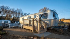A vintage camper with a metal staircase leading to the entrance, surrounded by other campers and trees.