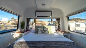 A narrow, well-lit interior with a bed, air conditioning unit, and a window offering a view of a residential area.