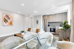 A modern living room with a glass dining table, white sofas, and a kitchen area with wooden cabinets and stainless steel appliances.
