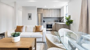 A modern living room with a sofa, coffee table, and glass dining table. A kitchen with appliances and cabinets is visible in the background. - Plateau Prime Residence (Montreal)