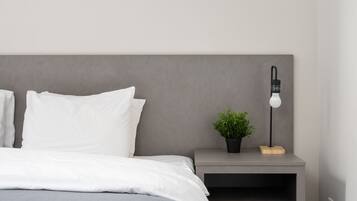 A modern bedroom with a grey headboard, white bedding, a bedside table with a lamp, and a small plant.