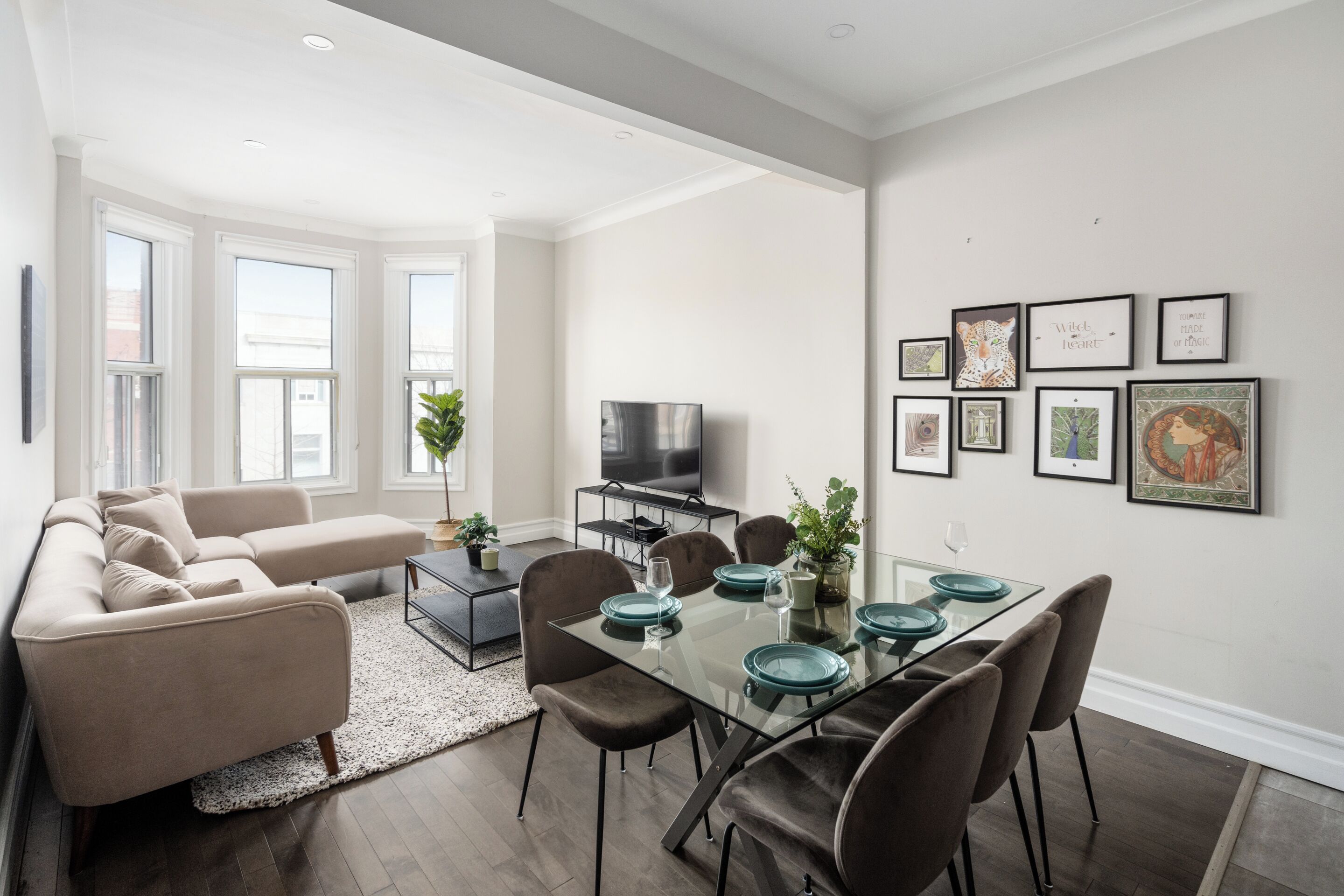 A modern living room with a glass dining table, brown leather chairs, a beige sofa, and a flat-screen TV.