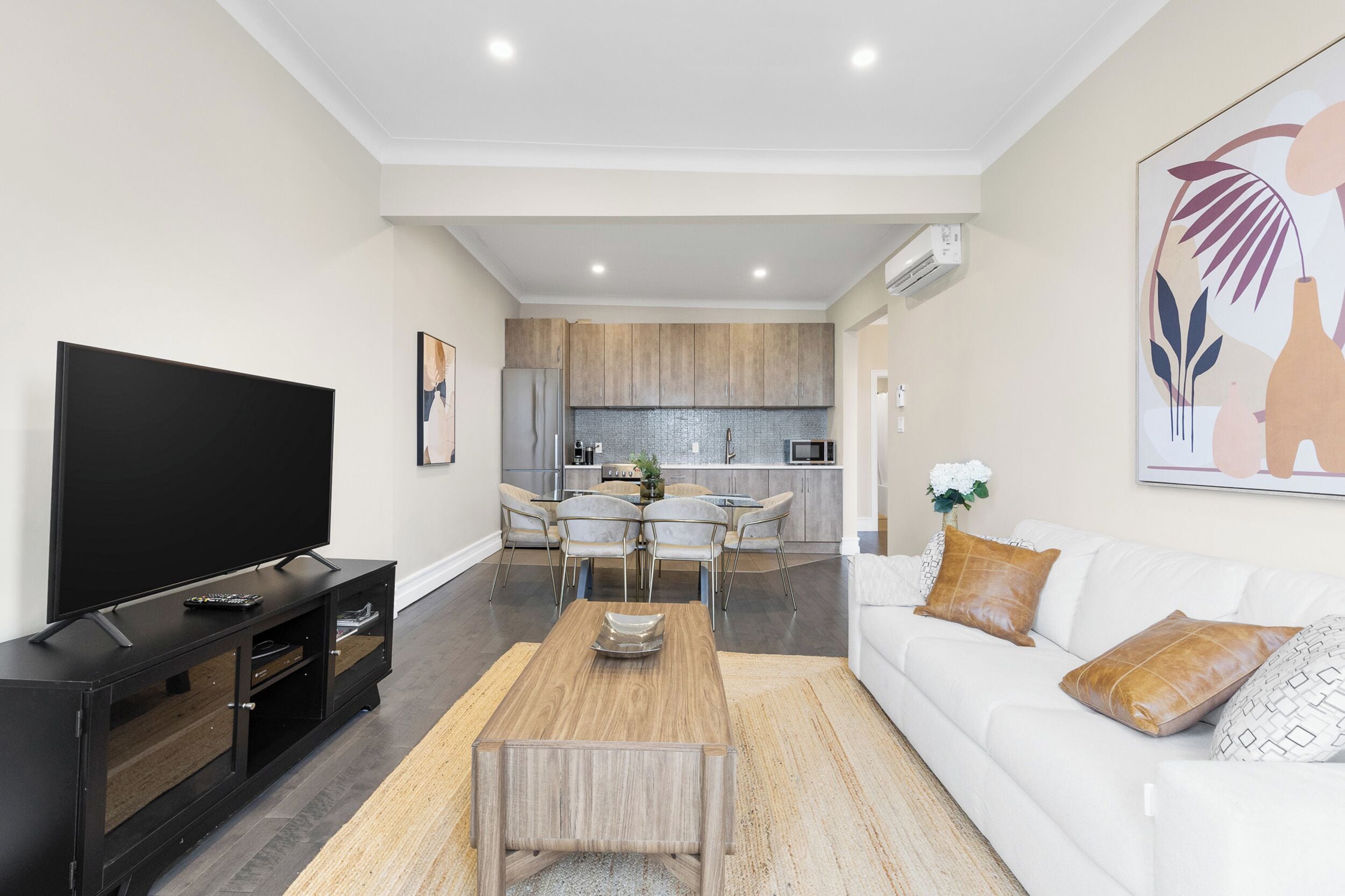 Una sala de estar moderna con un televisor de pantalla plana, una mesa de centro de madera y un sofá blanco con almohadones decorativos. Al fondo, se observa una zona de cocina con una mesa de comedor y sillas.