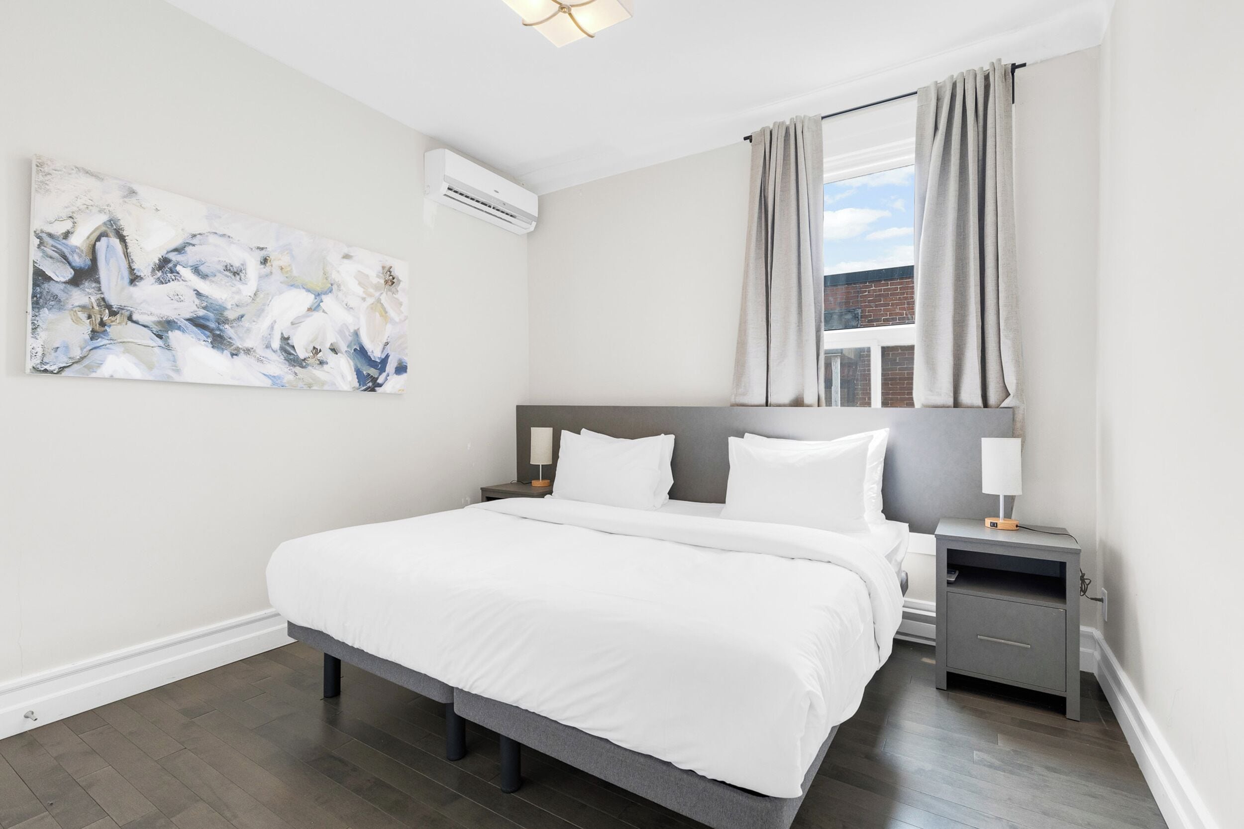 A modern bedroom with a large bed, a nightstand with a lamp, a wall-mounted air conditioner, and a window with curtains.
