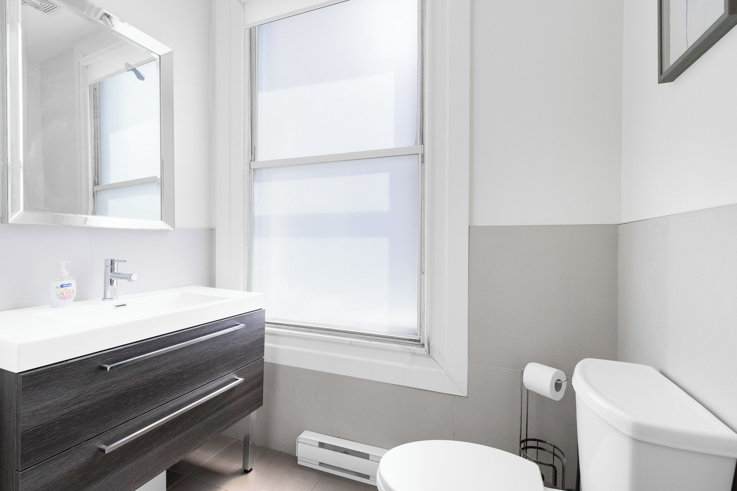 Un baño moderno con un lavamanos blanco, tocador de madera oscura, un ventanal grande y un inodoro.