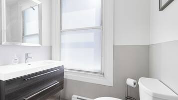 Un baño moderno con un lavamanos blanco, tocador de madera oscura, un ventanal grande y un inodoro.