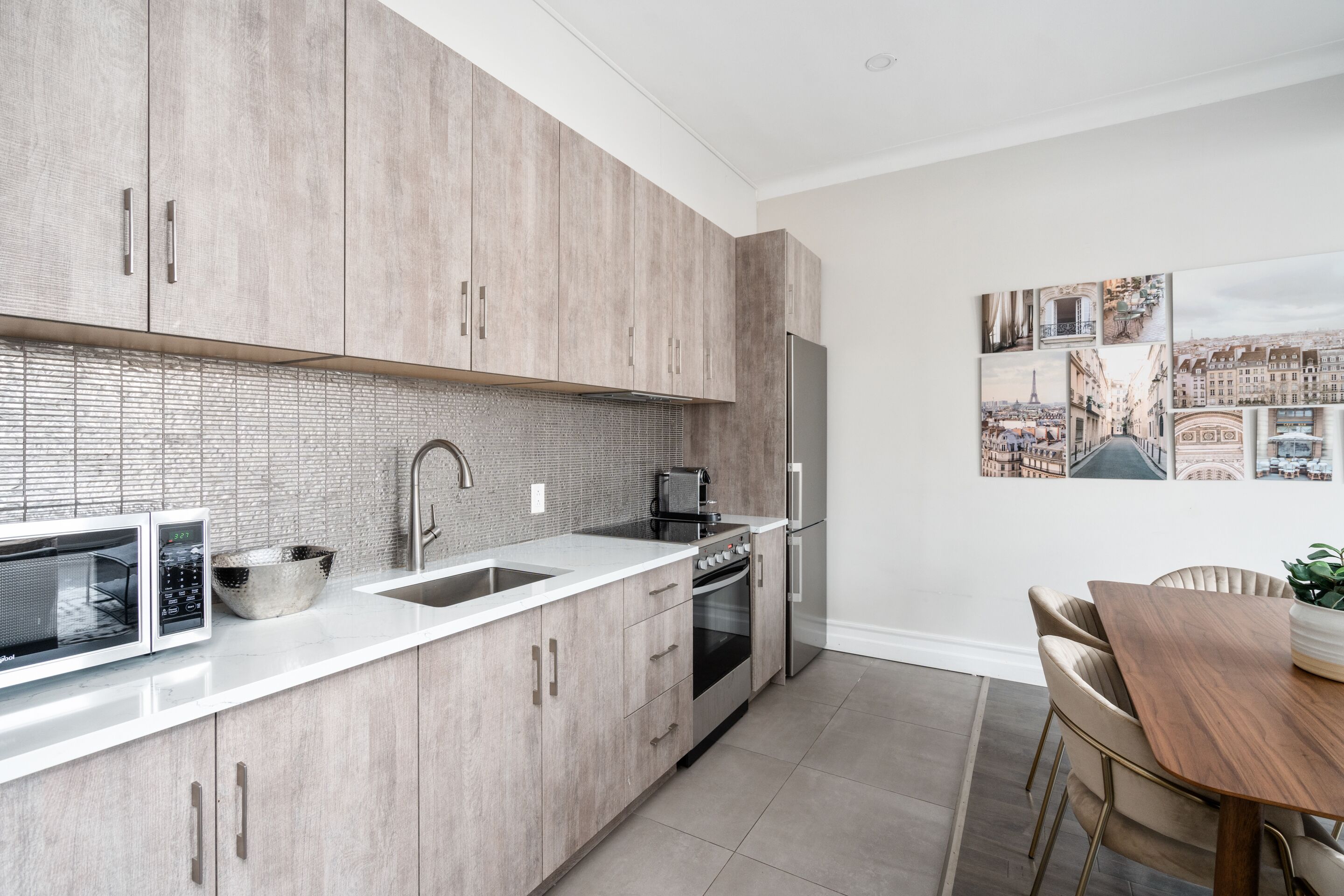 A modern kitchen with light wood cabinets, a white countertop, a sink, a stove, and a refrigerator. There is a dining table with chairs and a potted plant.