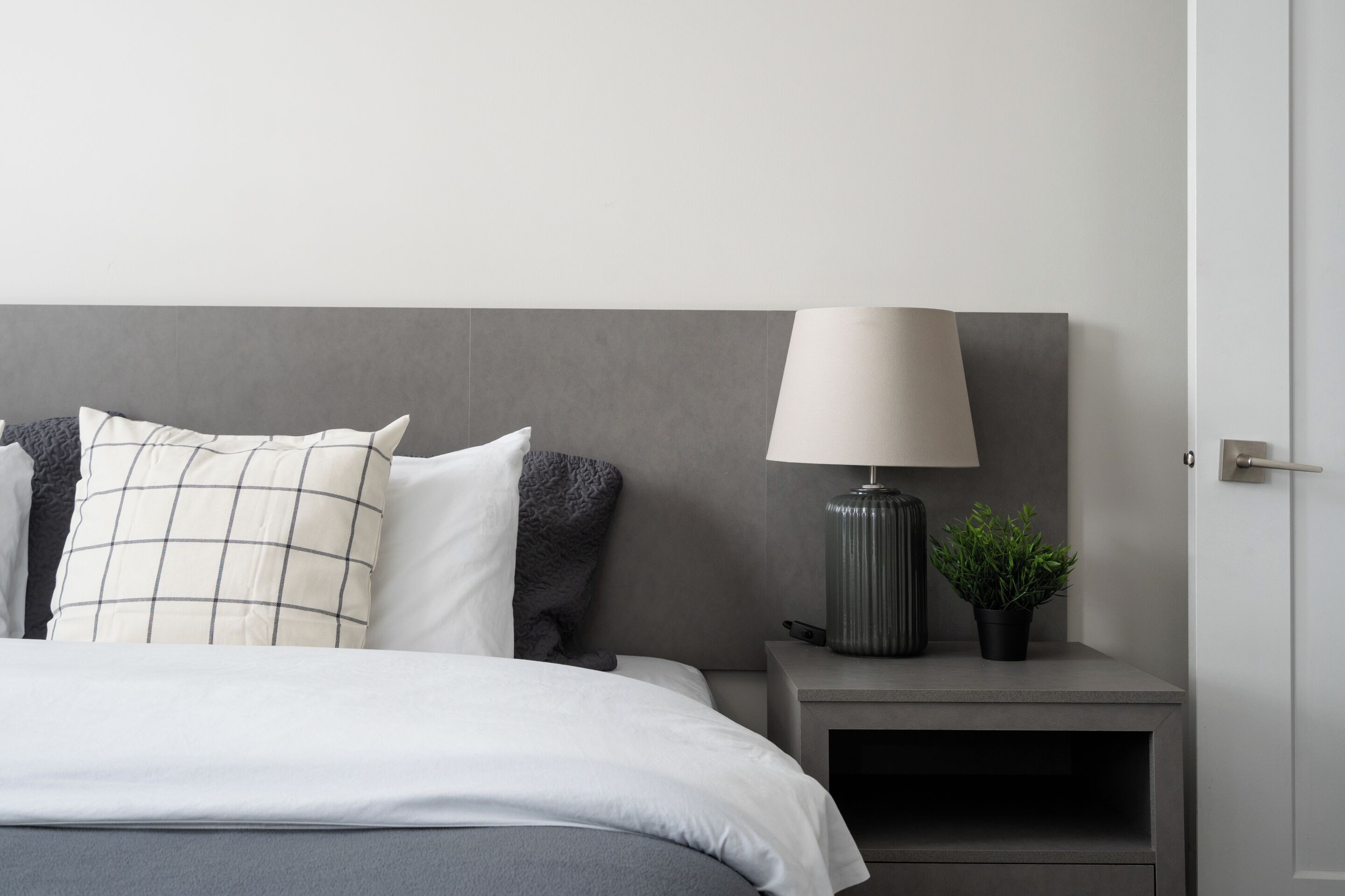 Ein ordentlich bezogenes Bett mit grauem Kopfteil, ein Nachttisch mit Lampe und einer kleinen Pflanze, und eine Tür rechts.