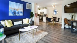 Una sala moderna con una pared azul decorativa, un sofá negro y una mesa de centro. Hay una zona de cocina con armarios blancos y una mesa de comedor con sillas.