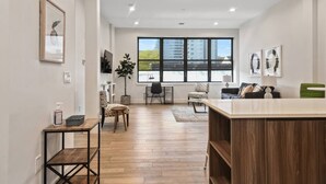 Een moderne woonkamer met een houten vloer, een groot raam, een groene bank en een eethoek met een tafel en stoelen.