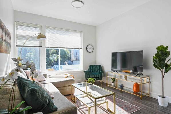 A modern living room with a sofa, a coffee table, a TV on a stand, and a large window with blinds.
