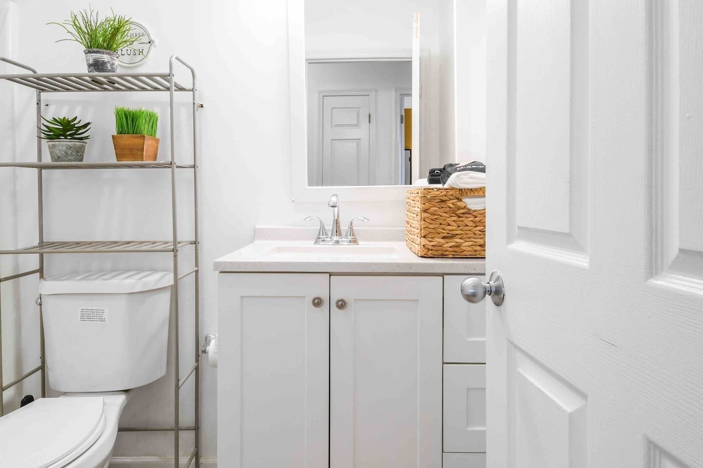 A small, white bathroom with a toilet, a sink, and a mirror. There are shelves with plants and a basket on the sink counter.